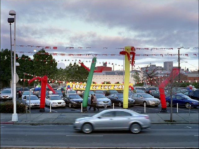 Video Reference: car lot, parked cars, inflatables, banner, street, moving car, outdoor, dealership, cloudy sky, urban, vehicles, parking lot, road, sale sign