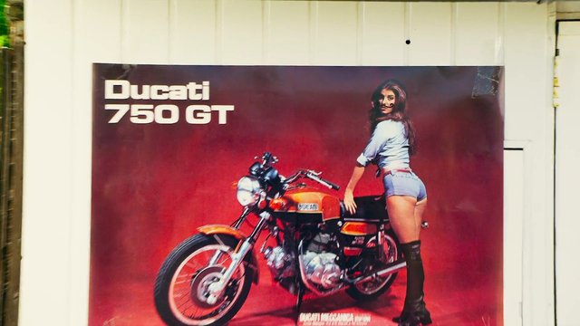 Video Reference: poster, motorcycle, Ducati, 750 GT, advertisement, woman, red background, wall, vintage bike, vehicle, portrait, garage, print