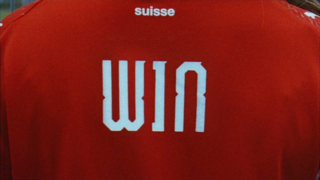 Video Reference: sports jersey, red shirt, back view, team uniform, text, close-up, fabric, athletic wear, suisse, white lettering