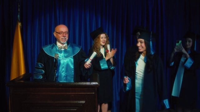 Video Reference: Smile, Outerwear, Microphone, Curtain, Gesture, Academic dress, Entertainment, Graduation, Scholar, Headgear