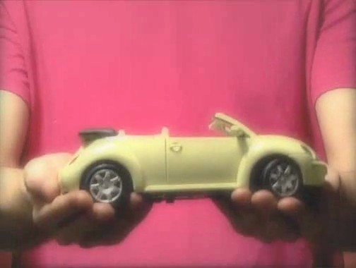 Video Reference: toy car, convertible, yellow, hands, pink shirt, close-up, model car, indoor, plaything, vehicle