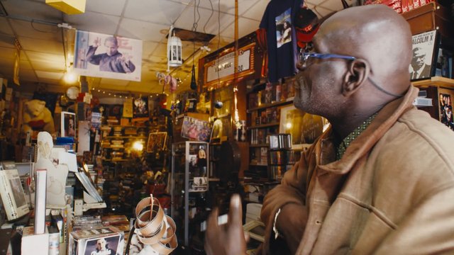 Video Reference: man, profile, indoor, shop, gallery, books, posters, shelves, cluttered, warm lighting, glasses, interior, retail, memorabilia, casual clothing