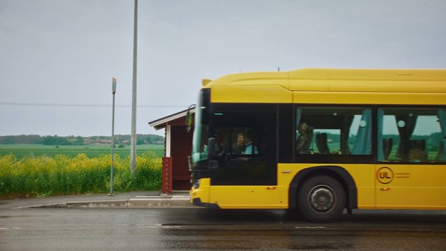 Video Reference: bus, yellow bus, road, wet pavement, rural, countryside, fields, roadside stop, cloudy sky, public transport, motion blur, bus stop, outdoors