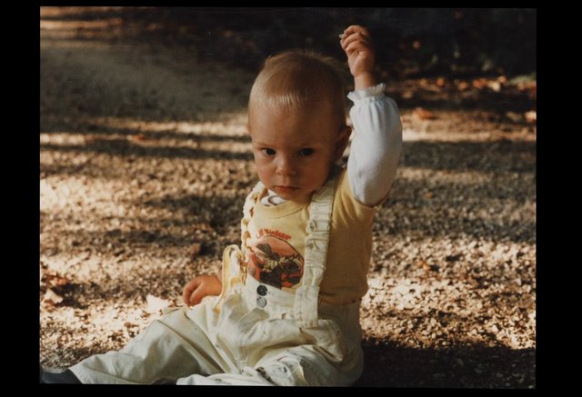Video Reference: child, toddler, outdoors, sitting, raised hand, sunlight, dappled light, portrait, playground, casual clothing, cute, human, ground, nature