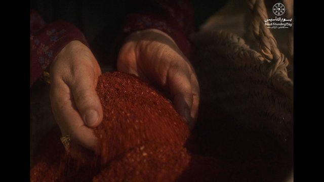 Video Reference: hands, red powder, granules, pouring, close-up, texture, warm light, dark background, festival logo, Saudi Founding Day, traditional clothing, craft, material, scooping, ritual, macro