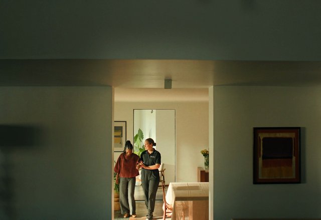 Video Reference: indoor, doorway, two women, walking, conversation, living room, framed art, chair, table, vase, flowers, home interior, soft light, domestic scene