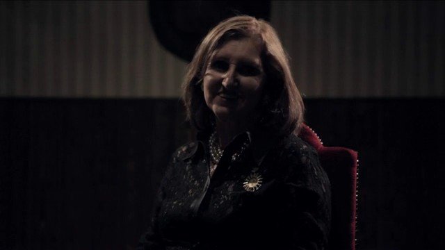 Video Reference: portrait, woman, indoor, low_light, seated, shadow, dark_background, chair, candid, interview, moody, backlit