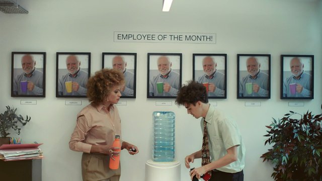 Video Reference: office, break room, employee of the month, portraits, water cooler, two people, indoor, wall display, workplace, drinks, plants, desk, documents, office decor, framed photos