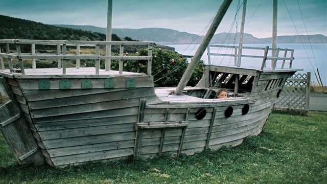 Video Reference: wooden boat, ship structure, grass, coastline, water, hills, outdoors, rustic, weathered wood, child, deck, railing, shoreline, sky, landscape, playground
