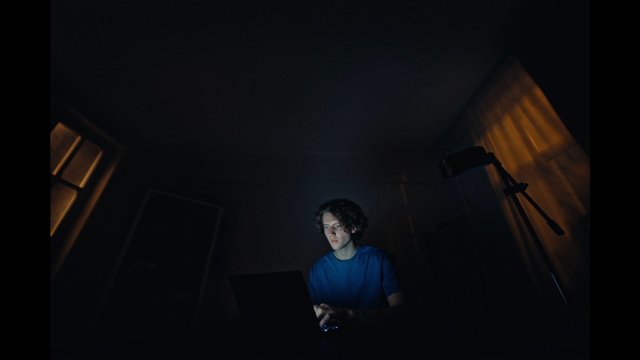 Video Reference: person, laptop, working, dark room, indoor, night, blue shirt, computer, low light, moody, home office, silhouette, warm light