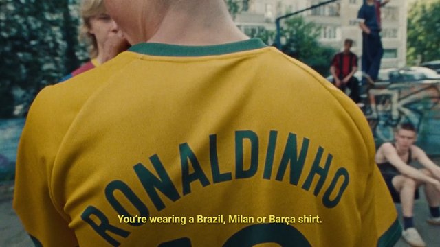 Video Reference: soccer, jersey, yellow shirt, green trim, outdoor, group of people, city courtyard, back view, subtitles, sportswear, crowd, buildings