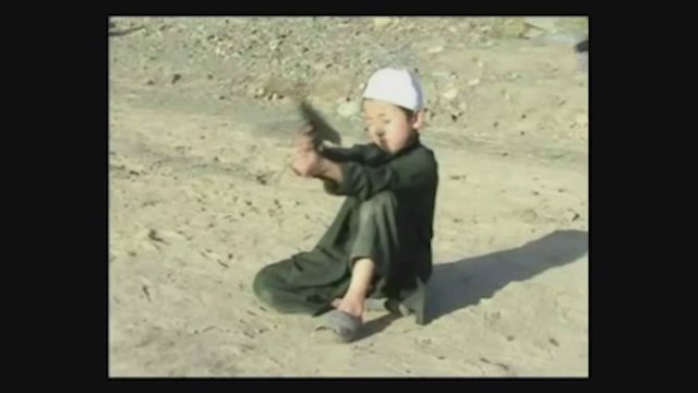 Video Reference: child, boy, sitting, outdoors, sand, white cap, dark clothing, hands, object, desert, play, action, seated, sunlight