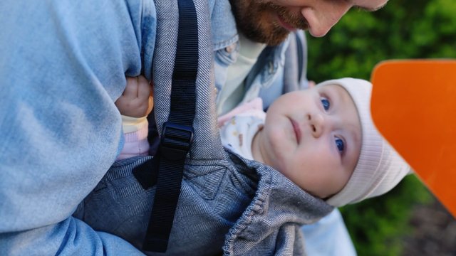 Video Reference: baby, infant, parent, carrier, outdoor, close-up, family, portrait, green background, blurred background, holding, sling, hood, soft focus
