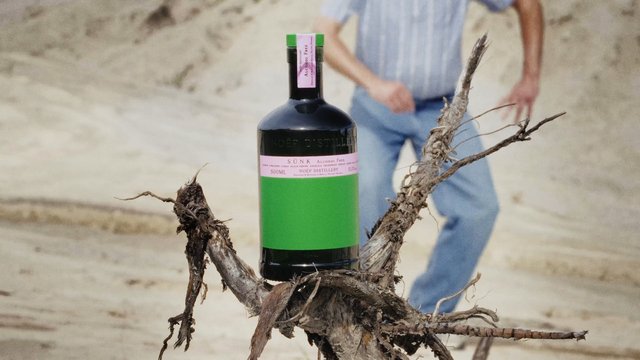 Video Reference: bottle, liquor, driftwood, sand, outdoors, beach, product, label, still life, person, blurred background, close-up