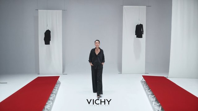 Video Reference: runway, fashion show, woman, black outfit, minimalist stage, white studio, red carpet, hanging garments, catwalk, model, modern set, brand text, event, presentation, studio lighting