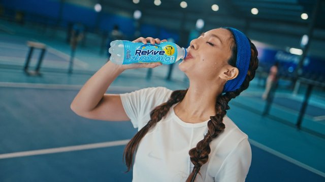 Video Reference: woman, sports drink, bottle, tennis court, indoor court, athlete, hydration, fitness, blue background, close-up, action, sportswear, summer, commercial, beverage