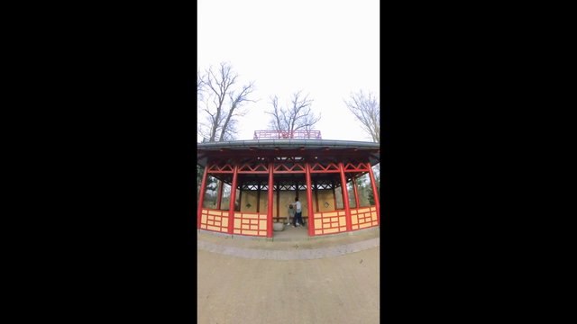 Video Reference: pavilion, gazebo, park, outdoor, architecture, red structure, yellow panels, trees, overcast sky, people, vertical video, tourism, shelter, walkway, public space