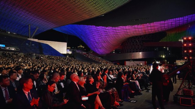 Video Reference: audience, applause, outdoor event, night, crowd, stage, venue, canopy, architecture, colorful lights, ceremony, concert, spectators, evening, formal attire