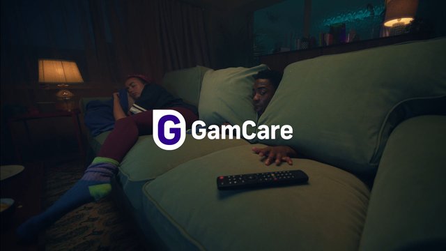 Video Reference: living room, couch, two people, remote control, dim lighting, evening, interior, relaxing, sofa, lamp, television, home, overlaid logo, advertisement, quiet scene