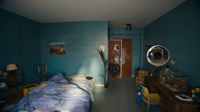 Video Reference: bedroom, interior, unmade bed, blue walls, person, doorway, boombox, fish tank, desk, chair, shelves, lamp, instrument, tuba, clutter, apartment