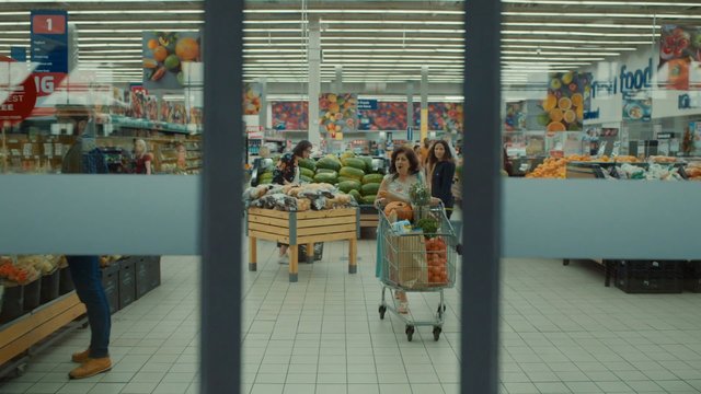 Video Reference: supermarket, grocery store, produce section, shopping cart, shoppers, fruits, vegetables, interior, retail, aisle, fresh produce, consumer, food market, glass doors, bright lighting