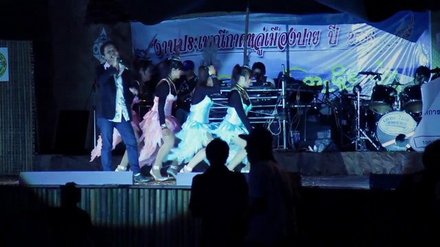 Video Reference: stage performance, live concert, singer, dancers, band, drum set, audience, night event, indoor stage, colorful costumes, music show, Thai banner, performance lighting, crowd silhouettes