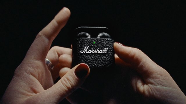 Video Reference: hands, earbuds, charging case, wireless earbuds, close-up, black device, green light, audio accessory, portable case, dark background, technology, brand logo