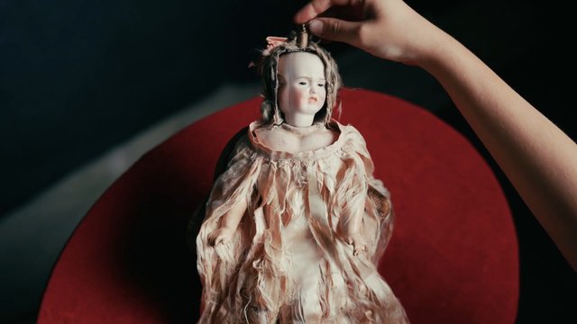 Video Reference: doll, toy, vintage, porcelain, dress, hand, adjusting, close-up, dark background, red circle, creepy, antique, figurine, suspense, portrait