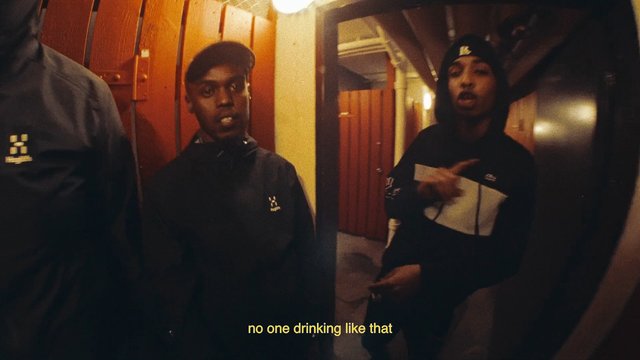 Video Reference: indoor, hallway, three men, hoodies, caps, gesturing, wooden doors, warm lighting, subtitles, group, standing, corridor, casual clothing, low light, video frame