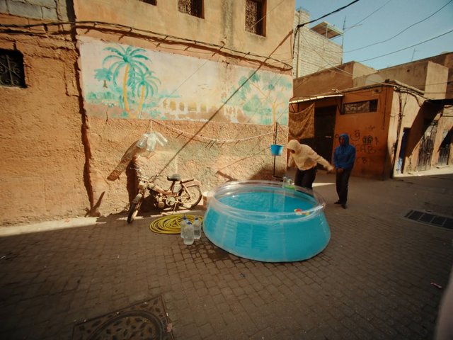 Video Reference: alley, street, buildings, mural, palm trees, motorcycle, plastic pool, water basin, people, hooded clothing, sunlight, urban, courtyard, clothesline, buckets, outdoor