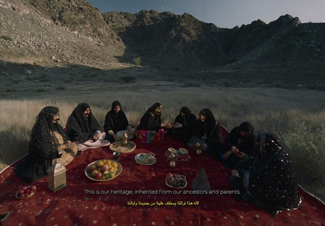 Video Reference: outdoor, women, group, traditional meal, picnic, carpet, mountains, valley, cultural gathering, food trays, desert, rural, ceremony, sharing, landscape, subtitles