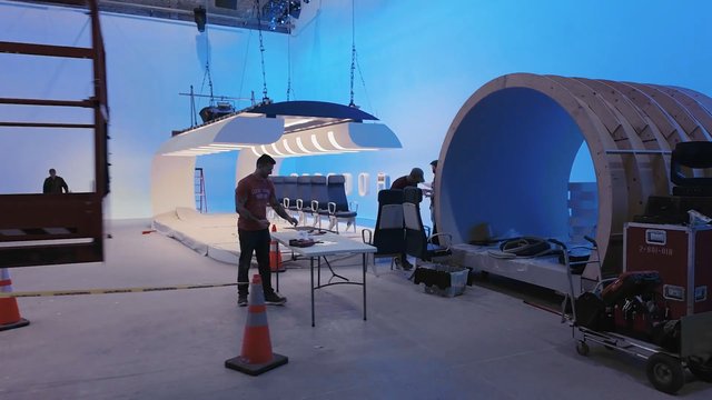 Video Reference: film set, production stage, studio, blue screen, airplane cabin, set construction, crew, lighting rig, chairs, equipment cart, indoor studio, behind the scenes, prop set, interior set