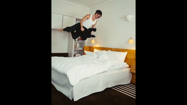 Video Reference: bedroom, man, jumping, bed, indoors, action, midair, white tank top, black pants, furniture, interior, motion, casual, home, playful