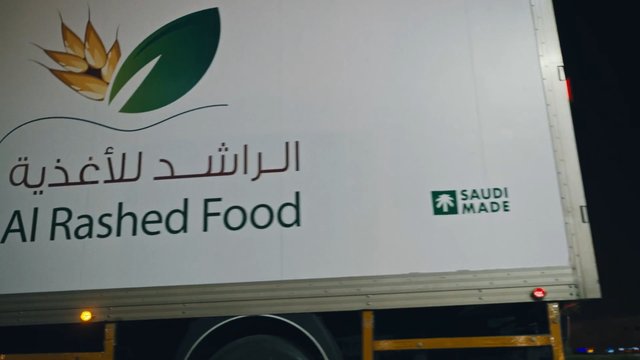 Video Reference: truck, trailer, delivery, food, branding, Arabic text, English text, vehicle side, logo, Saudi Made, night, transport, commercial vehicle
