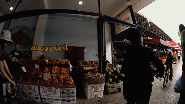 Video Reference: market, fruit stall, produce, bananas, apples, oranges, watermelons, shopping, shoppers, bicycle, indoor market, covered walkway, vendor, stall, fresh fruit, aisle