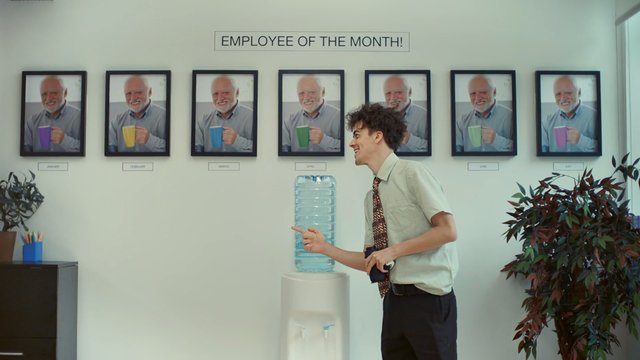 Video Reference: office, employee of the month, wall display, framed photos, water cooler, man, dress shirt, tie, break room, plants, indoor, corporate, portrait photos, standing, pointing, workspace