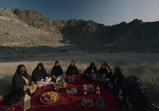 Video Reference: women, outdoor, mountain, valley, carpet, group, traditional clothing, fruit, dishes, gathering, desert landscape, rocky terrain, seated, ceremony, rural, picnic