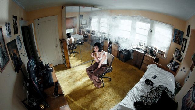 Video Reference: bedroom, person, chair, smoke, mirror, desk, computer, posters, bed, window, indoors, cluttered, cigarette, vapor, home, interior