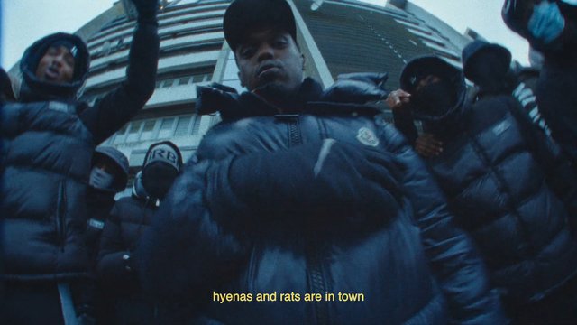 Video Reference: group, people, hoodies, puffer jackets, face coverings, urban, high-rise building, low angle, street, outdoors, crowd, subtitles, winter clothing, dramatic, music video