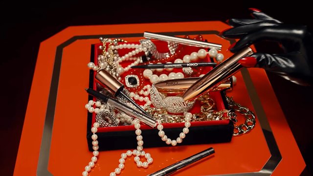 Video Reference: makeup, cosmetics, lipstick, eyeliner, mascara, jewelry, pearls, gloves, close-up, still life, red box, orange surface, beauty products, hands, dark background