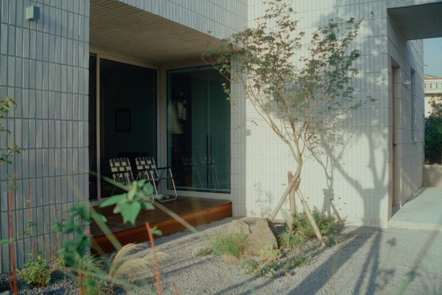 Video Reference: house, exterior, modern architecture, tiled wall, entryway, garden, tree, landscaping, doorway, patio, chairs, sunlight, residential, minimalist, gravel, shadows
