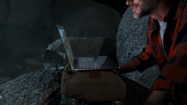 Video Reference: man, laptop, outdoors, night, rock, hiking, backpack, plaid shirt, technology, remote work, camping, dark scene