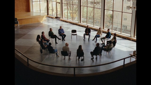 Video Reference: group, people, seated, circle, meeting, discussion, indoor, stage, hall, windows, sunlight, chairs, interior, community, seminar, assembly
