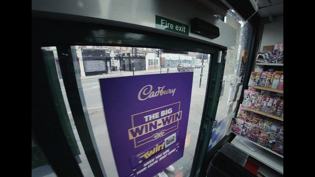 Video Reference: shop interior, glass door, fire exit, street view, Cadbury poster, magazine rack, convenience store, newsstand, retail, urban, window display, poster, doorway, sidewalk, magazines