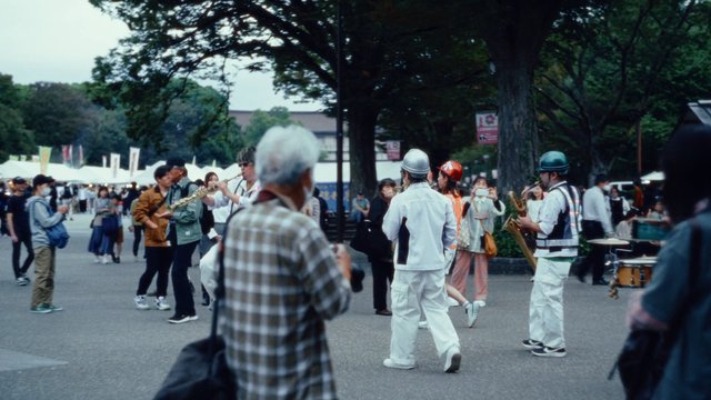 Video Reference: outdoor, public gathering, park, crowd, street performance, brass band, musicians, spectators, festival, trees, walking, tourism, daytime, live music