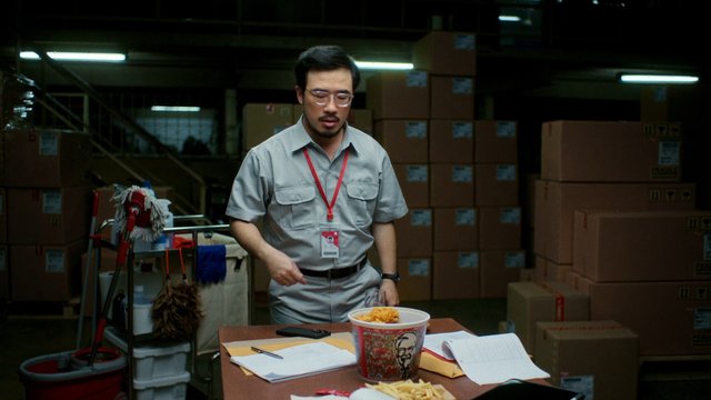 Video Reference: man, warehouse, storage room, cardboard boxes, work uniform, lanyard, papers, fast food, table, indoor, dim lighting, cleaning tools, office