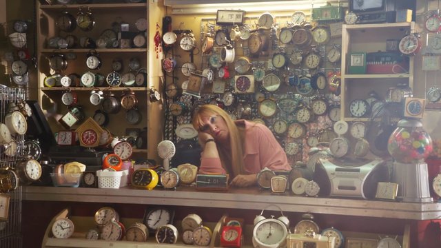 Video Reference: clock shop, alarm clocks, wall clocks, shop interior, counter, woman, sitting, shelves, display rack, vintage clocks, timepieces, cluttered, indoor, retail, small business