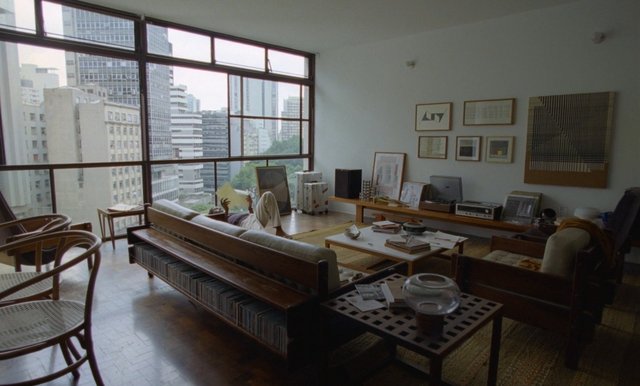 Video Reference: living room, city view, large windows, modern interior, sofa, coffee table, wall art, apartment, books, chairs, person lying down, wooden floor, urban skyline, minimalist decor, daylight, interior design