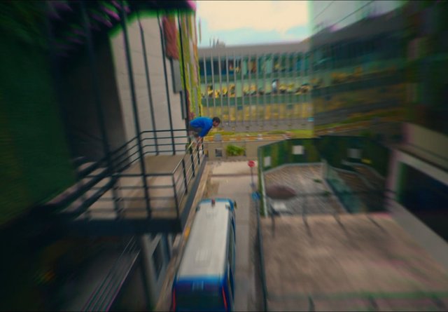 Video Reference: urban, building, ledge, person, blue jacket, crouching, street, bus, railings, alley, city, motion blur, outdoor, height, architecture