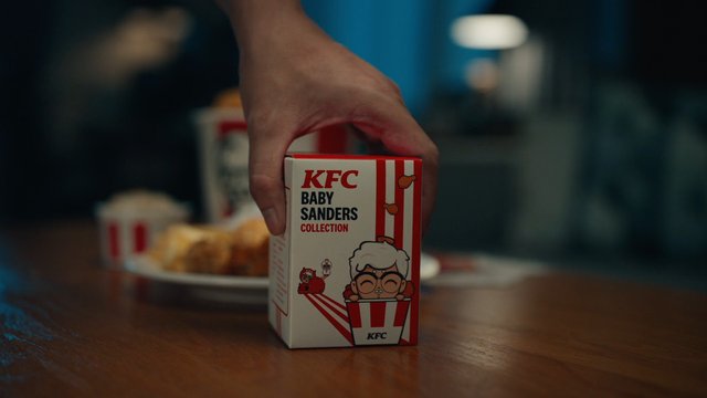 Video Reference: hand, box, collectible, fast food, fried chicken, table, package, KFC, toy, product shot, indoor, close-up, brand packaging, restaurant, blurred background